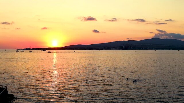 Sunset casts warm hues over water as boats sail peacefully, framed by trees and distant hills, creating a tranquil coastal atmosphere