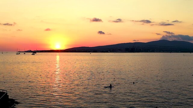 Sunset casts warm hues over water as boats sail peacefully, framed by trees and distant hills, creating a tranquil coastal atmosphere