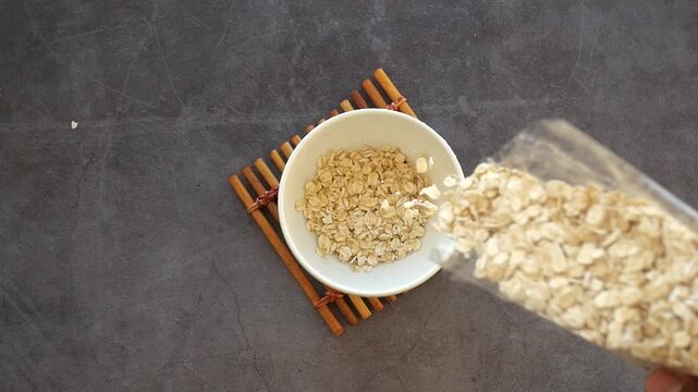 Slow motion video of rolled oats falling from plastic package into white ceramic bowl on bamboo mat