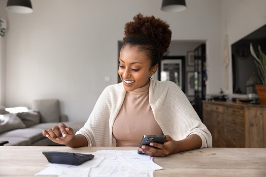 Happy African woman calculates personal finances at home using calculator and smart phone, seated at table with bills and receipts. Budgeting, financial literacy, remote work, and digital lifestyle
