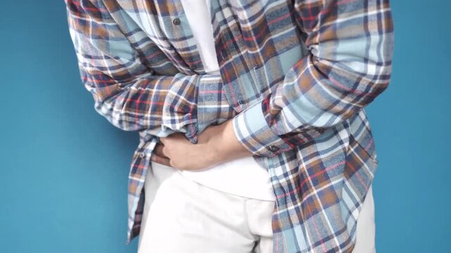 A man in a casual plaid shirt and white t-shirt clutches his abdomen in discomfort, experiencing stomach pain or indigestion, captured in a close-up dynamic shot against a blue background