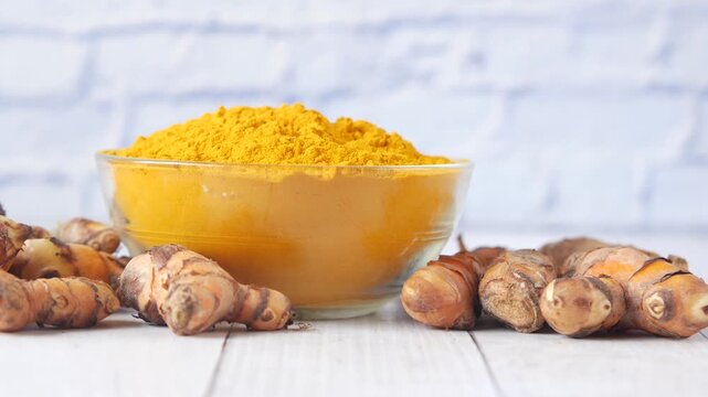 Ground turmeric powder in glass bowl with fresh whole roots on white wooden table against brick wall background