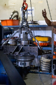 Heavy planetary gear assembly suspended by chain hoist in mechanical repair workshop.