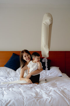 Asian mother and toddler son celebrating first birthday on white bed with cake and balloon