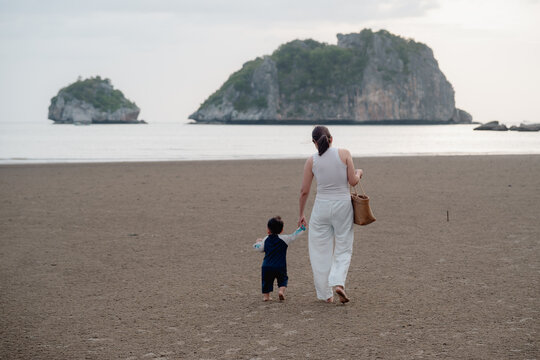 Asian mother holding hands with toddler son walking on sandy beach together with sunset.