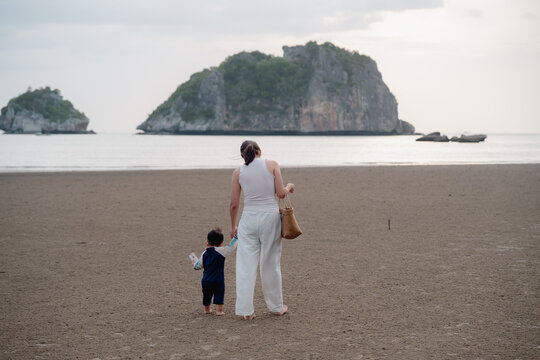 Asian mother holding hands with toddler son walking on sandy beach together with sunset.