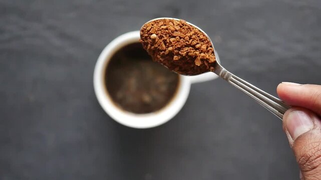 Person pouring instant coffee granules from silver spoon into white ceramic cup on dark surface top view