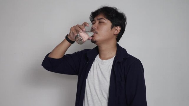 Asian man drinking water from transparent glass tumbler isolated on plain white background, healthy lifestyle hydration concept, quenching thirst, daily water intake habit, and health care routine.