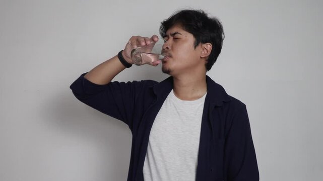Asian man drinking water from transparent glass tumbler isolated on plain white background, healthy lifestyle hydration concept, quenching thirst, daily water intake habit, and health care routine.