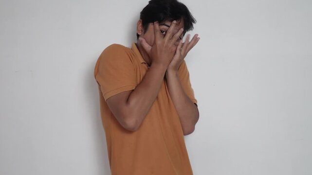 Scared Asian man in orange shirt covering his face with hands and peeking through fingers in fear, frightened, horror reaction, and phobia concept against white background.
