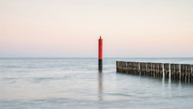 Red beacon and wooden breakwater in the calm sea during a serene sunset. Perfect for maritime background and travel concepts.