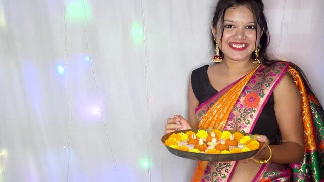Two Indian women sharing traditional sweets kaju katli holding a golden platter filled with assorted colorful flower and sweets, celebrating a Diwali festival.