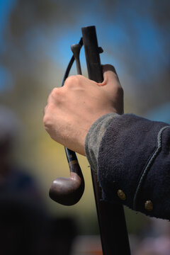 Close-up of a Civil War reenactor gripping a period-style long gun while a wooden tobacco pipe hangs alongside it.