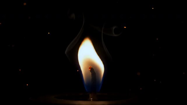 Candle flame flickers in total darkness with tiny floating embers nearby.