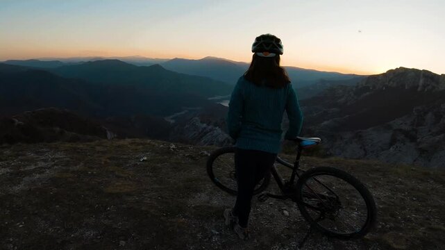 Woman cyclist standing with mountain bike on cliff edge overlooking sunset landscape.