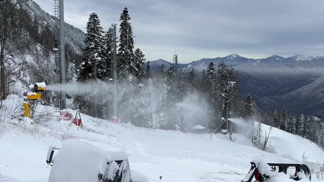 Snowmaking machine operates in a winter landscape. Snow-covered ground and evergreen trees surround the area. Mountains are visible in the background.
