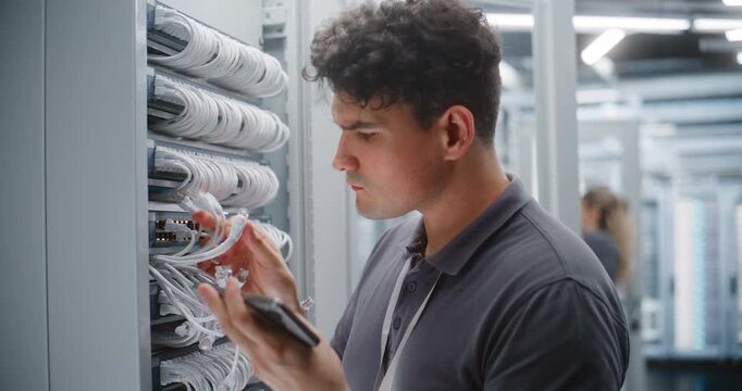 Male Data Center Technician Verifies Ethernet Patching and Physical Port Labels Against Network Documentation on smartphone. Female Colleague Performs Maintenance on Adjacent Server Rack in Facility.