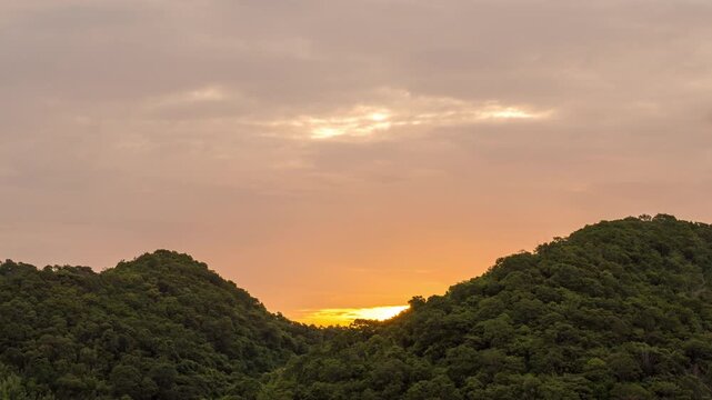 Time lapse view of a beautiful golden sunrise behind a lush green mountain range. Vibrant cloudy sky with orange and yellow morning sunlight over the forest landscape.