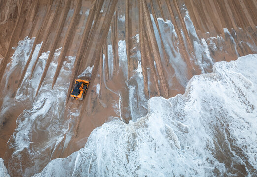 A tracked earthmover works along a wet sandy shoreline as ocean waves wash across freshly graded sections of beach. Parallel machine tracks and reshaped sand indicate active beach maintenance or coast