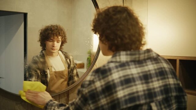 Person cleans mirror in bathroom during daytime with yellow cloth while wearing checkered shirt and apron, showing focus on the task at hand