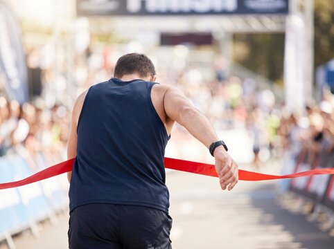 Back view of a male athlete crossing the finish line and breaking the red tape ribbon at an outdoor marathon competition with spectators cheering.