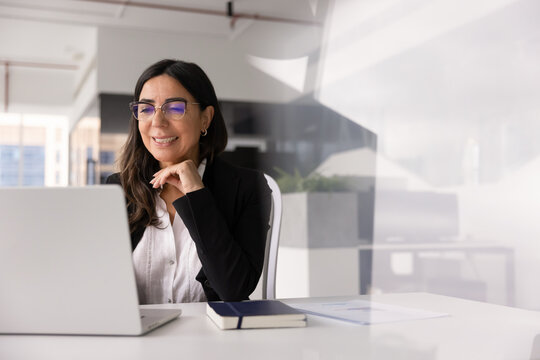 Smiling middle-age businesswoman working on laptop in office, reviewing positive business report results, replying on email, represents successful, effective workflow, corporate tasks management at