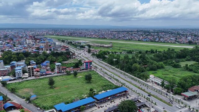 High angle aerial view of a long straight divided highway separating empty green plots from a sprawling Nepalese city and distant airport runway