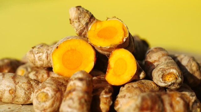 Close-up cinematic video of fresh yellow turmeric roots and slices on a wooden surface against a vibrant yellow background, capturing the natural texture and bright color with gentle sunlight