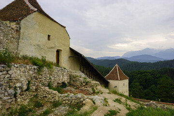 The Rasnov Fortress, a medieval citadel in Romania, stands against a backdrop of lush green forests and distant mountains under a cloudy sky. © Nick