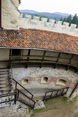 View of the courtyard of Bran Castle, Romania, with its stone walls, wooden stairs, and tiled roof, set against a backdrop of forested mountains. © Nick
