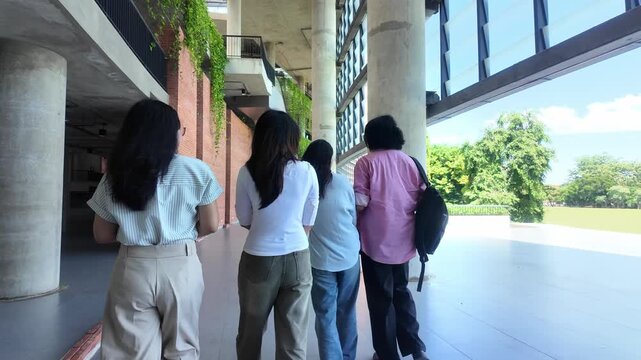 Group of young university student walking together away from camera through modern campus building corridor, college friends with backpack walking down university hallway and talking during break time