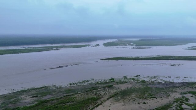Wide drone shot of a massive muddy river navigating around flat green sandbars and islands under a gloomy monsoon sky