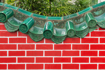 Detail of Jade green glazed tiles on top of a curved red brick wall with tropical plants behind © JPK