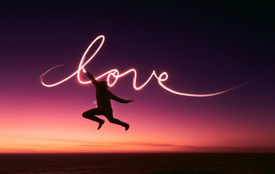 Silhouette of a woman jumping with the word love in light trail in sunset background with copy space