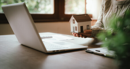 Fototapeta premium Real estate professional holding a model house at a desk with property documents, representing home ownership, mortgage, investment, and property transactions.