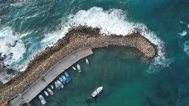 Drone aerial of overhead drone perspective shows fishing boats moored in a calm harbor protected by a stone pier from crashing turquoise ocean waves. Agios Georgios Peyia, Cyprus