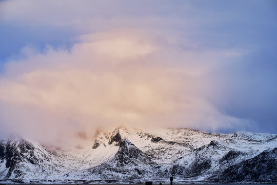 Majestic snow-capped mountain peaks covered in thick soft clouds during winter sunrise in Lofoten Islands, Northern Norway. Dramatic Arctic alpine landscape with pastel glowing clouds over rugged moun