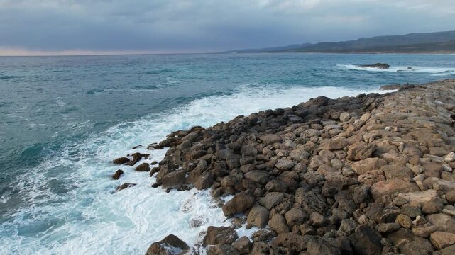 Drone aerial 4k ultra high definition Choppy sea waves with white sea foam break onto a rugged, rocky shore beneath dark, overcast storm clouds in the distance. Akamas Penisnula, Cyprus