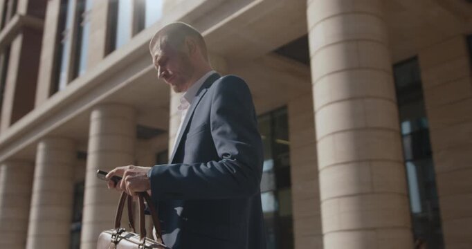 Camera circles a man in a blue suit checking his watch and smartphone while carrying a leather bag through a developing urban area with modern architecture and ongoing construction.