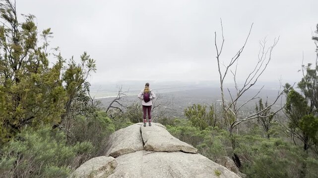 A girl hiker stands on a rock, admiring the beautiful view of the bush and plains at You Yangs Regional Park near Melbourne, Victoria, Australia.