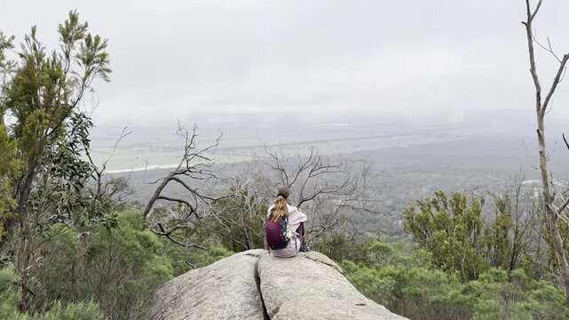 A girl hiker sits on a rock, admiring the beautiful view of the bush and plains at You Yangs Regional Park near Melbourne, Victoria, Australia.