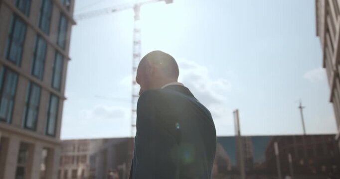Dynamic orbiting camera shot moves around a suited businessman using a mobile phone in a modern urban area with new development and construction cranes in the background.