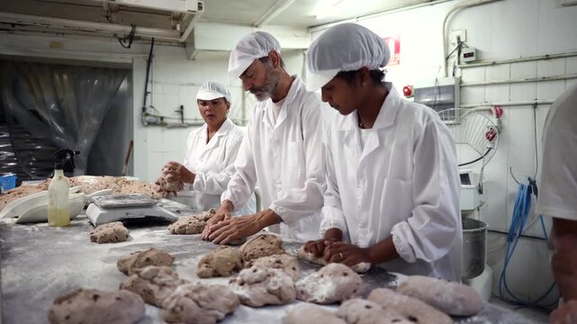 Team of dedicated bakers wearing white uniforms working together, preparing and shaping bread dough with raisins on a floured stainless steel table in a professional bakery production facility