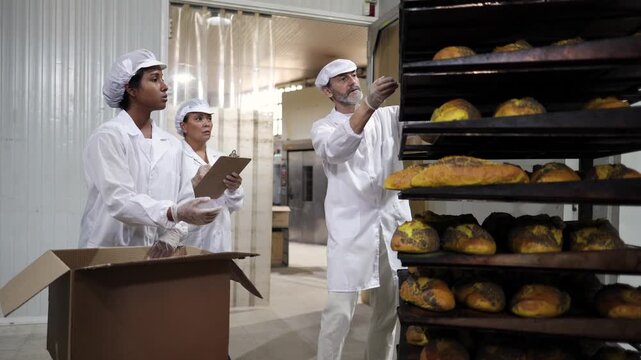 Diverse team of food industry workers in white coats and hairnets packing freshly baked loaves of bread into cardboard boxes for shipping, ensuring quality control in a commercial bakery