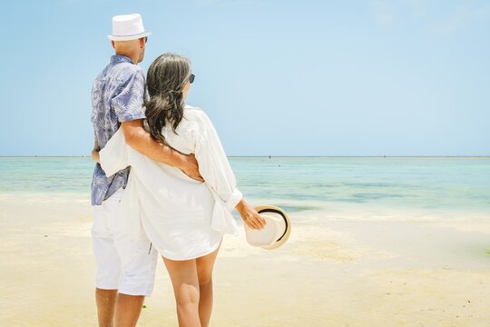 Couple stand together hugging hug enjoy arms up on white sand beach in sunny tropical destination. Romantic summer holiday by turquoise ocean with clear blue sky. Honeymoon paradise vibes.Zanzibar