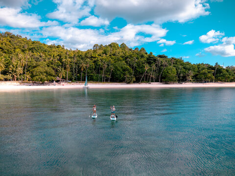 Top aerial view of couple of tourist are enjoy on water sport paddle sup-board with kayaking along at the edge of the sea beach under crystal clear of water in the island of summer trip