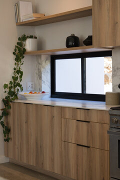 Light wood lower cabinets are framing white countertop under sliding frosted window with fruit bowl