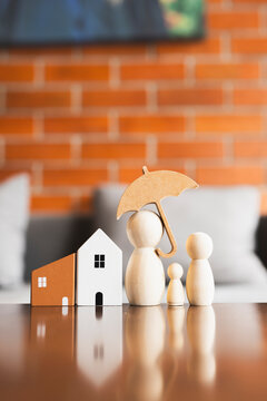 Family and home insurance concept with wooden peg doll figures under umbrella beside miniature house models on table with brick background for property mortgage protection and security.