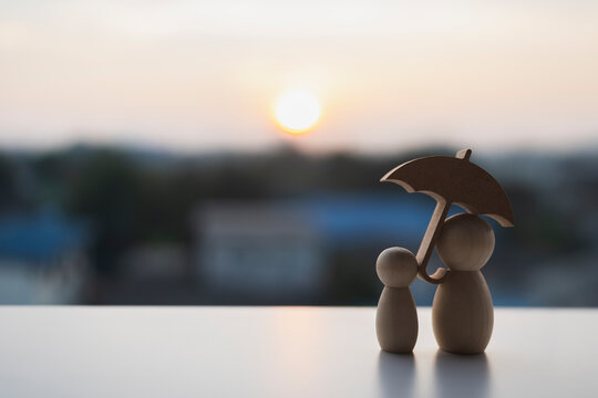 Single parent and child protection concept with wooden peg doll figures under umbrella on table against golden sunset background for family life insurance, custody care and future safety.