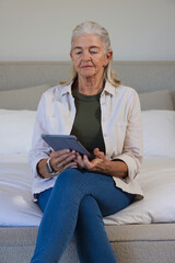 Senior woman holding gray tablet while reading on bed with pillows, wearing jeans and smartwatch © WavebreakMediaMicro